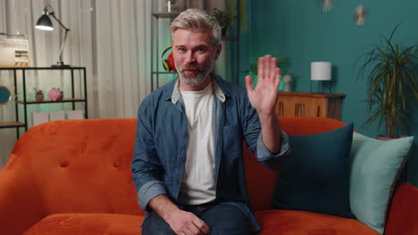 mature man smiling friendly at camera, waving hands gesturing hello, hi, greeting at home on couch