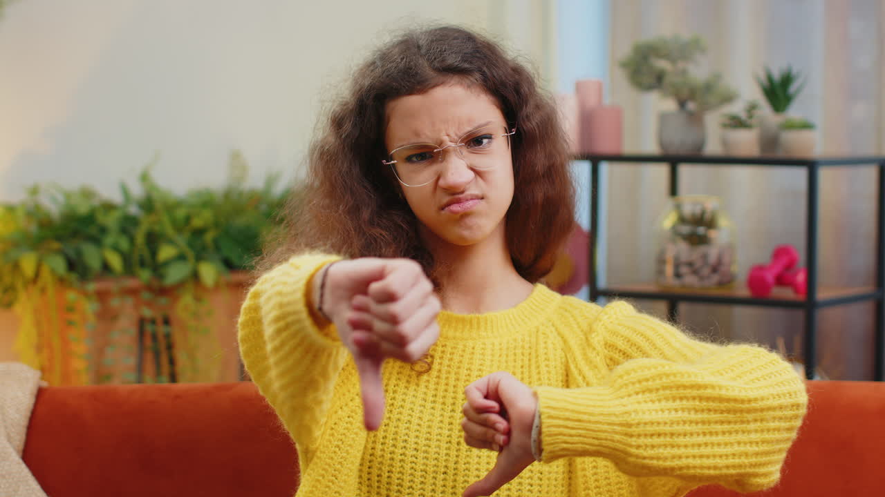 Upset child girl showing thumbs down dislike bad work disapproval dissatisfied feedback at home