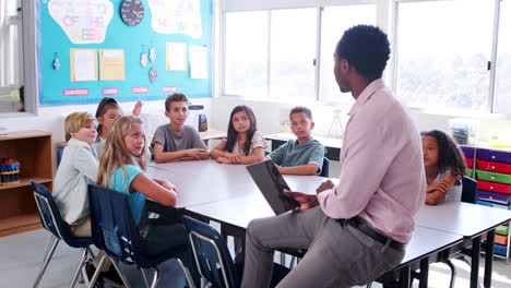 Teacher-sitting-desk-holding-tablet-computer-in-lesson