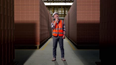 full body of asian male engineer with safety helmet thinking and looking around then raising his index finger while standing with red brick packed in stacks are stored