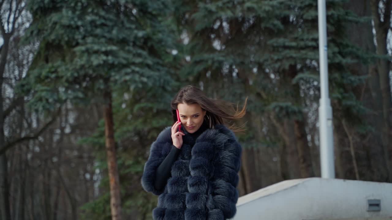 Luxury girl with a phone in the park. Portrait of attractive lady in fur coat walking in the park and talking the mobile phone. Slow motion.