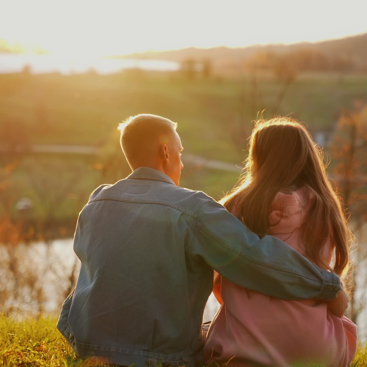 Romantic couple rests in nature. Young people boy and girl sitting together on grass and looking at sunset. Couple in love on date outdoors