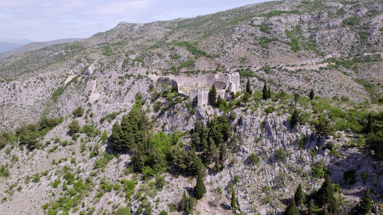 Le rovine della Fortezza di Blagaj si ergono tra creste rocciose e boschi verdi in Bosnia ed Erzegovina, rivelate da una ripresa aerea in ascesa che si inclina verso il basso.