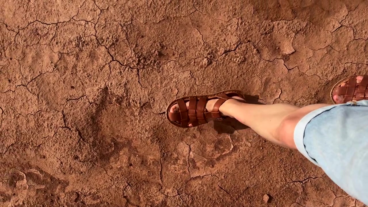 Persona che cammina su una superficie di terra secca e screpolata indossando sandali in un paesaggio arido
