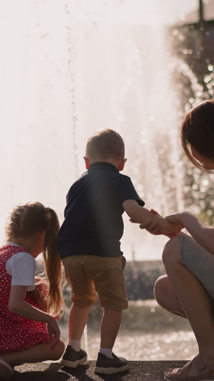 Mother with little son and daughter sits on parapet surrounding large fountain in park backside view slow motion. Woman and children refresh with splashing water drops