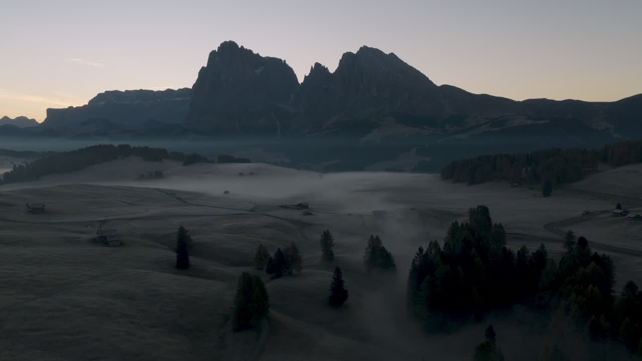 Filmisk luftfoto af frostklædte Alpe di Siusi-enge med spredte bjerghytter og morgentåge, dramatiske Dolomitter-toppe i de italienske alper ved solopgang, fredfyldt natursceneri