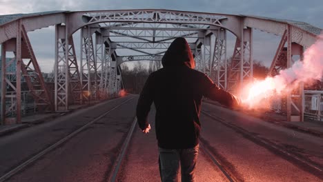 young man in hoodie and balaclava with red burning signal flare on the road under an old steel frame bridge, slow motion