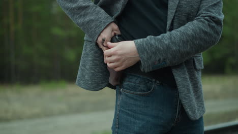 a close view of a man removing a concealed firearm under his jacket, standing beside a railway track, with blur forest view