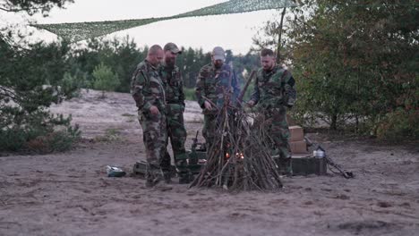 cuatro hombres en uniformes de camuflaje están de pie alrededor de una chimenea con una hoguera, fritando salchichas en palos, hombres uniformados están descansando después del servicio, ejercicios de campo