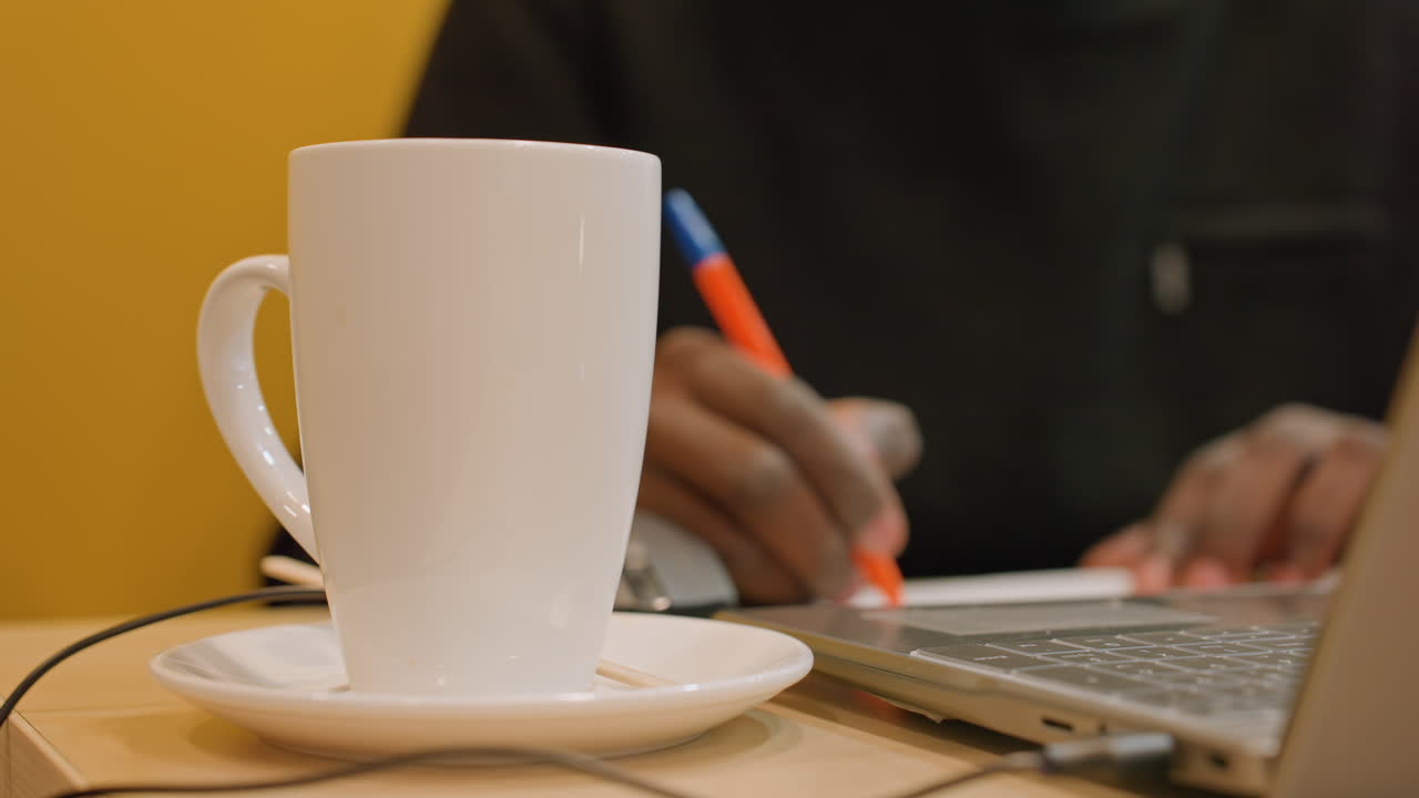 Tazza di caffè in primo piano, mani che scrivono accanto al laptop, scena di uno spazio di lavoro rilassato con tazza sul piattino, sfondo giallo caldo, routine mattutina multitasking, atmosfera accogliente e morbida profondità di campo ridotta