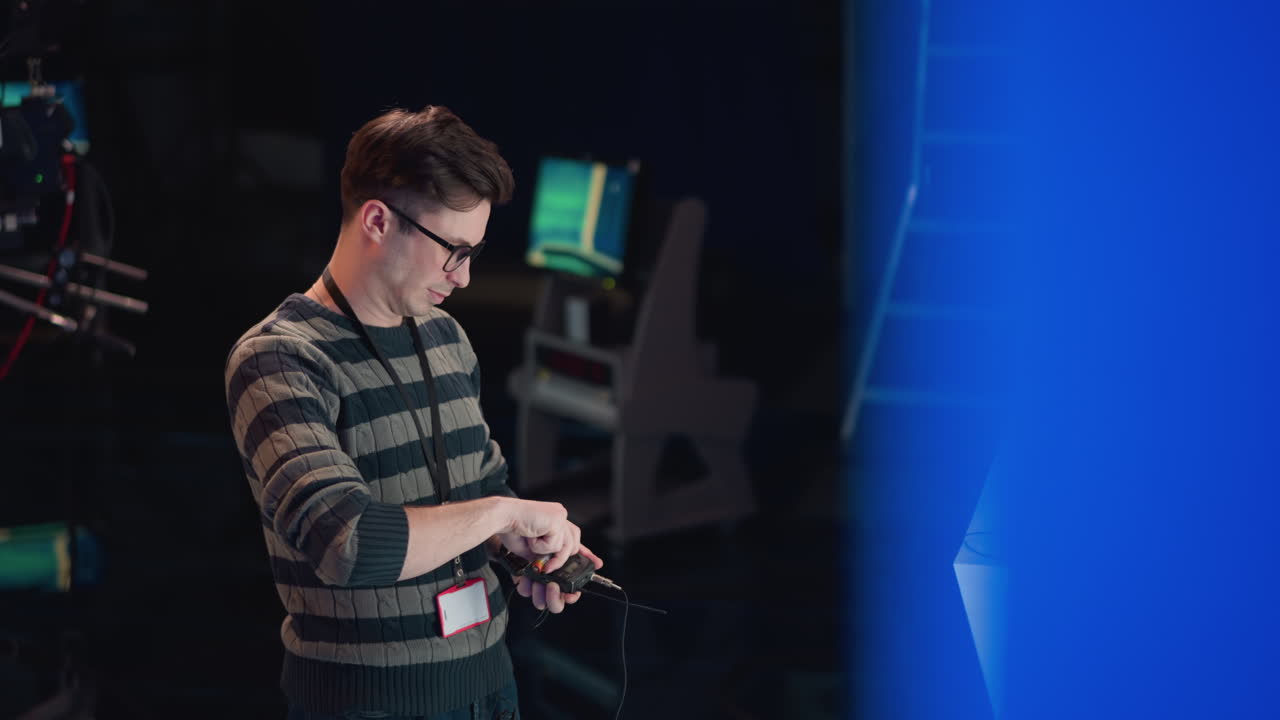 man wearing striped sweater and glasses adjusts wireless audio transmitter during video production setup indoors, with ladder, monitor and device on table, dimly lit studio prep scene