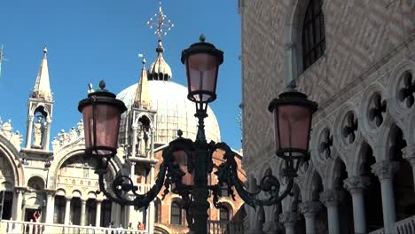 Tres-Farolas-En-Venecia,-Italia,-Con-La-Basílica-De-San-Marcos-Y-El-Palacio-Ducal-Al-Fondo-Mientras-Una-Paloma-Vuela-Hacia-Arriba.