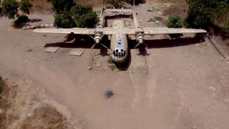 aerial turning and rising shot of an old dismantled army plane monument placed on an empty dirt field, surrounded by trees and bushes