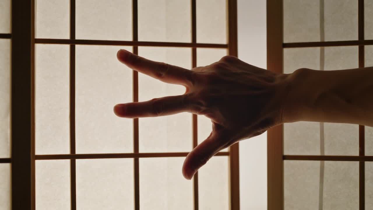 Mano destra in movimento con braccio scoperto che entra da destra attraverso uno shoji, dita allargate controluce, spazio per il testo.