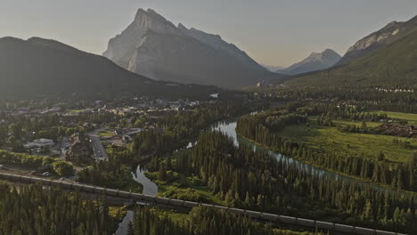 banff ab canada aerial v32 flyover forested valley capturing quaint town along bow river and views of rundle mountain ranges with train riding across the scene - shot with mavic 3 pro cine - july 2023