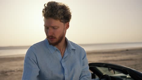 a bearded man with curly hair wearing a blue shirt and standing next to a black convertible, rolls up his sleeve and looks away against the yellow sky on the beach