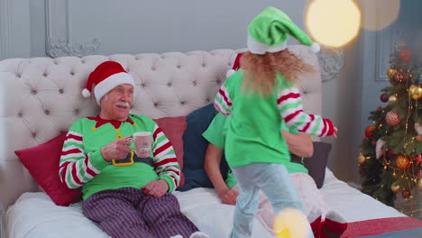 Senior-couple-grandparents-drinking-hot-tea-on-bed-with-granddaughter-toddler-kid,-Christmas-morning