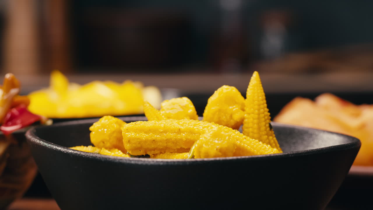 Close-up of fermented yellow mini corn with black pepper on black plate. Preservation of vegetables in glass jars. Fermentation preserved mini corns with spices.