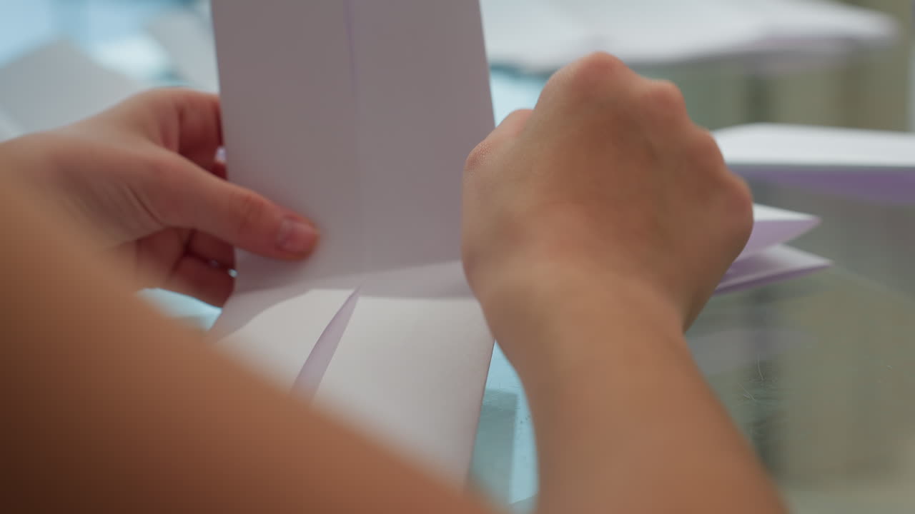 Hand view of child carefully folding paper one by one on the table, engaging in a creative activity, focus on hands and paper as they carefully fold the sheets in a home setting