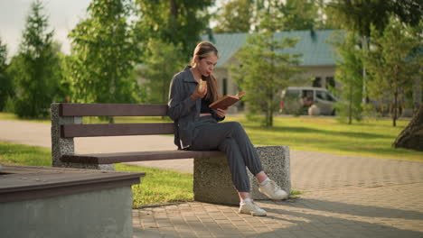 dama comiendo un bocadillo mientras lee un libro al aire libre, sentada en un banco del parque con la pierna cruzada, rodeada de vegetación, árboles y un edificio en el fondo