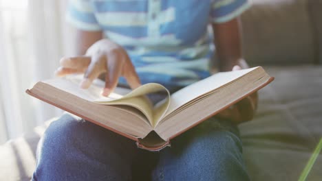 sección media de un niño afroamericano leyendo un libro sentado en el sofá en casa