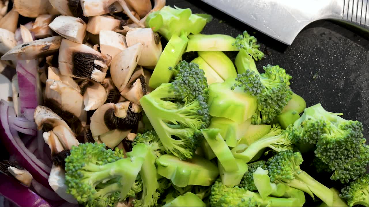 Close-up view of freshly chopped broccoli, mushrooms, and red onions are prepped on a dark cutting board. A chef's knife rests nearby, ready for healthy cooking and meal preparation