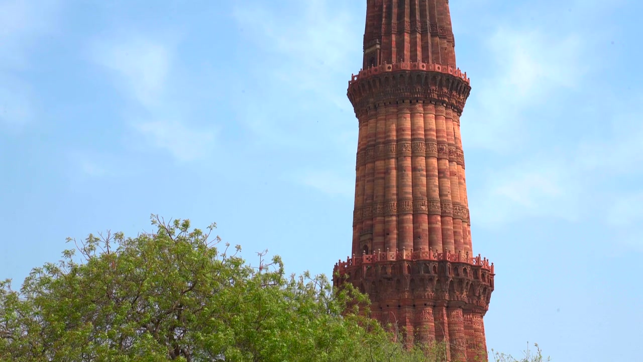 Kamera miring ke bawah menyorot struktur batu pasir merah detail menara Qutub Minar menuju pepohonan hijau di New Delhi, India