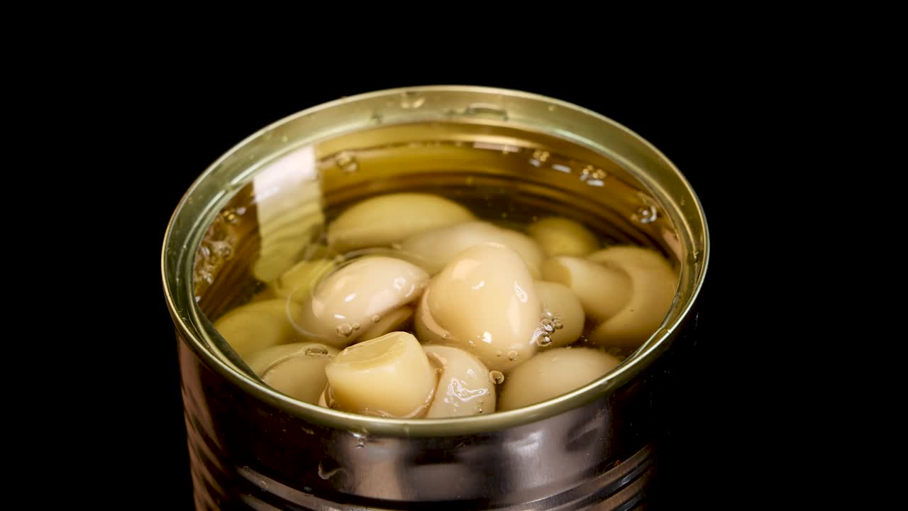 A stream of water splashes into an open can of champignon mushrooms, creating bubbles and ripples under bright studio lighting against a black background
