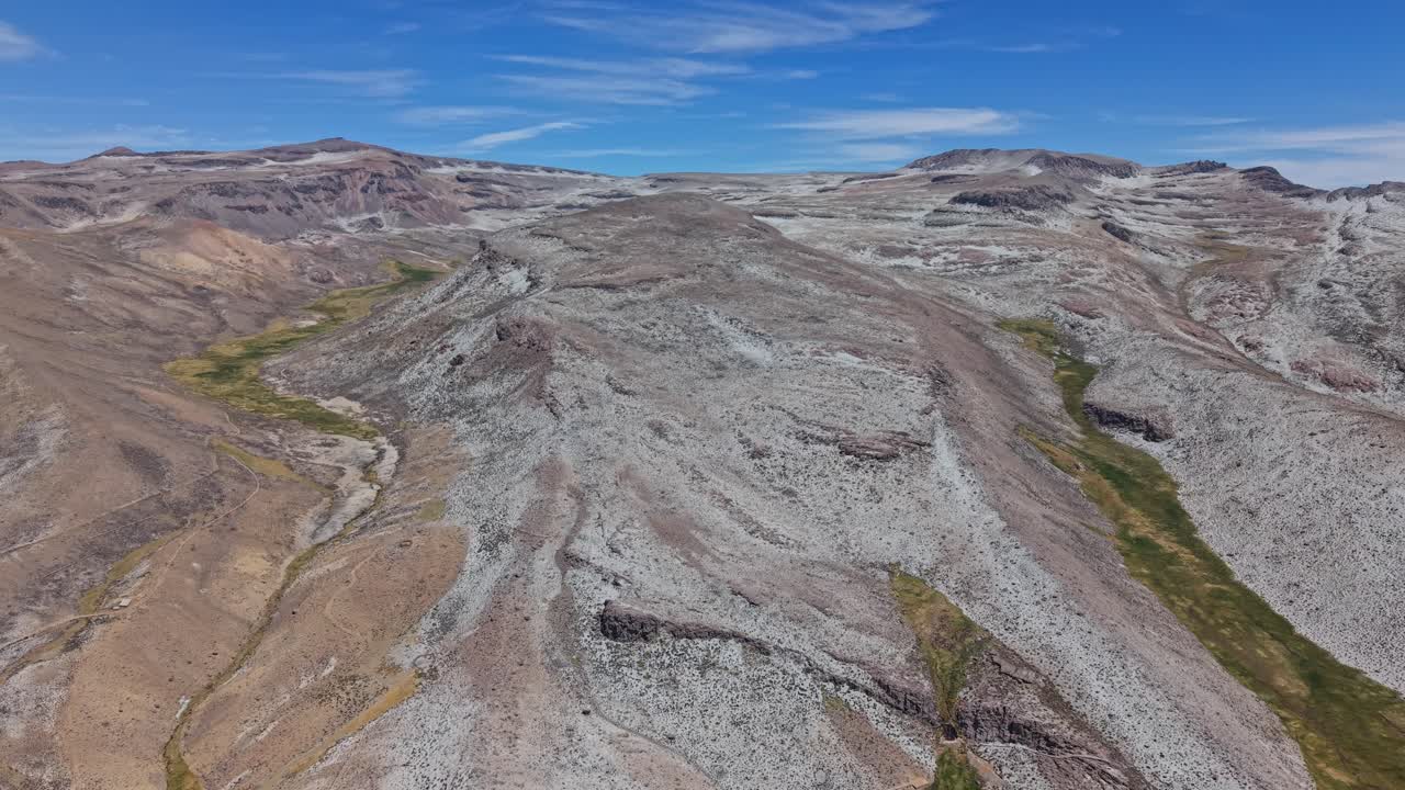 Um drone aéreo acompanha uma paisagem vulcânica acidentada, com picos de montanhas esculpidos pela erosão e cobertos de sal branco da Laguna Salinas. Manchas de enxofre e depósitos minerais revelam texturas geológicas impressionantes