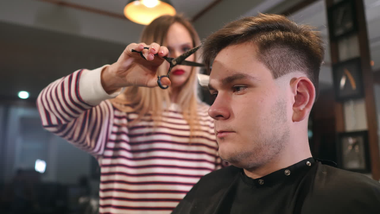 Interior shot of working process in modern barbershop. Side view portrait of attractive young man getting trendy haircut. Male hairdresser serving client, making haircut using metal scissors and comb.