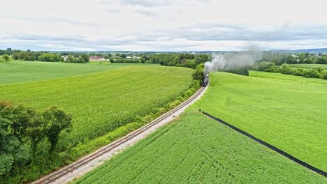 Eine-Luftaufnahme-Von-Amish-Ackerland-Mit-Einem-Einzigen-Eisenbahngleis-Und-Einem-Dampf-Personenzug,-Der-Sich-Nähert