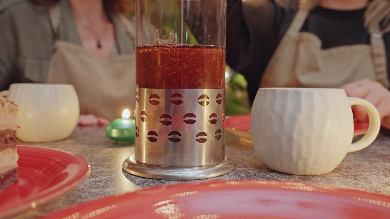 Fiorista che condivide un pasticcino con l'apprendista, tè caldo in infusione in una pressa di vetro, tazza in ceramica e torta a strati su un piatto rosso, candela e piccolo ornamento verde, scaffali del negozio sullo sfondo con vasi e piante.