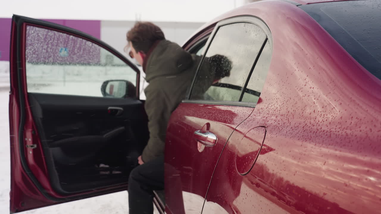 young adult in winter jacket steps out of red car and shuts door in snowy environment with blurred view of industrial building and leafless tree in background