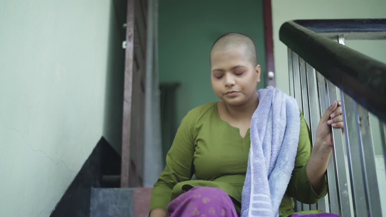 Giovane ragazza indiana calva, debole e malata che soffre di cancro, è stanca e siede sulle scale ansimando