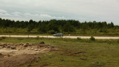 aerial side shot following a grey car in a open dusty straight road near some trees in a cloudy day