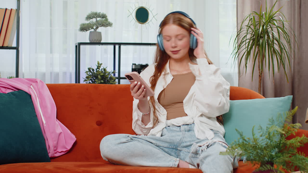 무선 헤드폰을 입은 행복한 십대 소녀가 집에서 소파에 누워서 좋아하는 음악을 듣고 휴식을 취하고 있습니다.