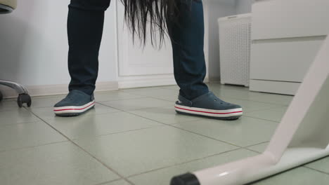 specialist wearing clinical crocs stands beside spa bed as long dark hair strands fall from client, tiled floor, swivel chair caster and grey storage basket visible under bright overhead lighting
