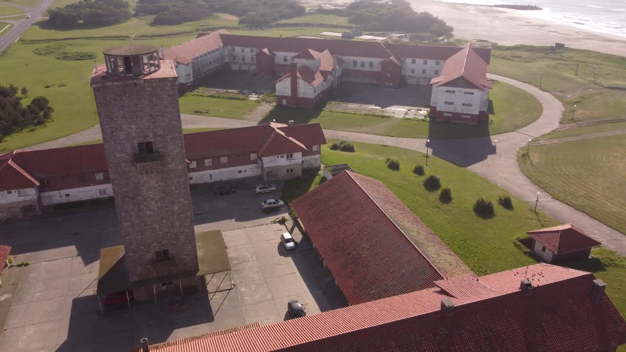 Aerial orbit shot of old farming hotel with tower and historic buildings beside beautiful sandy beach and ocean during summer - Chapadmalal in Argentina