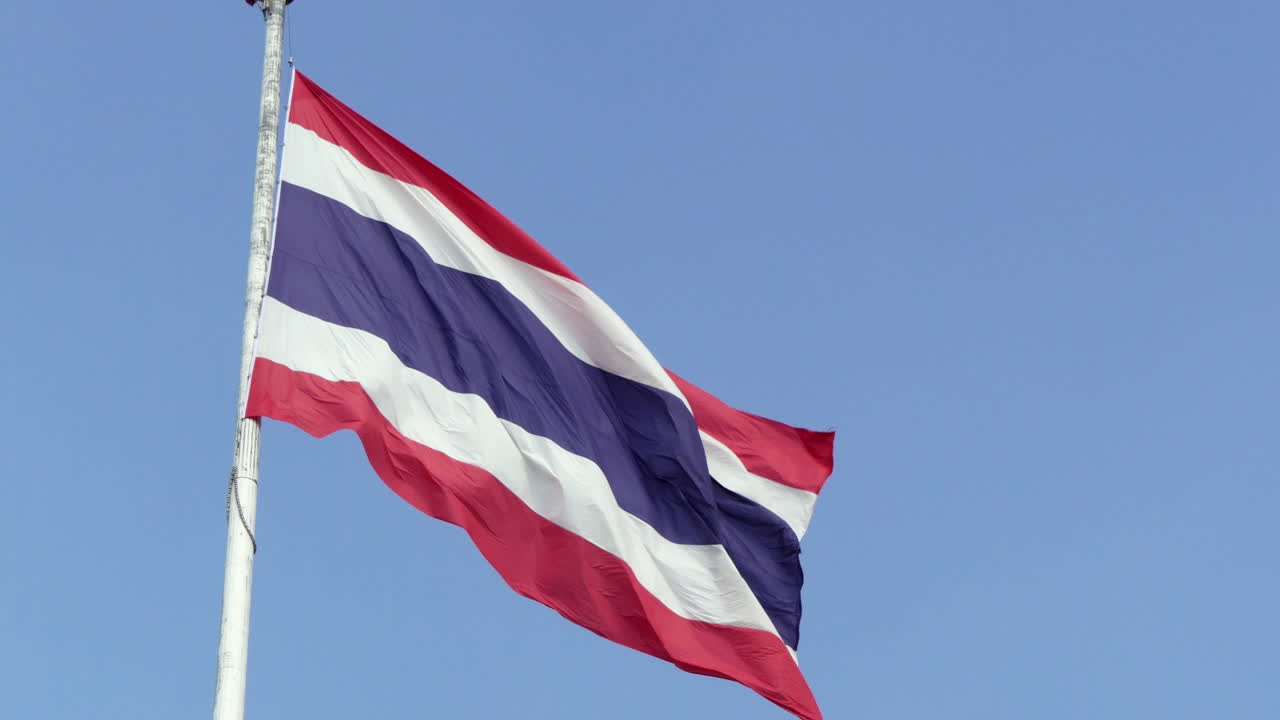 A majestic slow-motion shot captures the Thai flag fluttering at the top of a flagpole in the breeze, symbolizing the country's love and pride for its heritage.