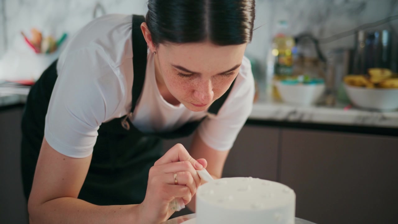 Giovane donna si concentra intensamente mentre decora una torta bianca. Si china vicino, mostrando concentrazione e cura nel suo lavoro. Lo sfondo della cucina include ciotole di frutta e utensili da forno.