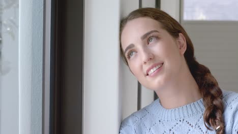 Thoughtful-caucasian-woman-touching-her-face-and-smiling-at-window-in-slow-motion