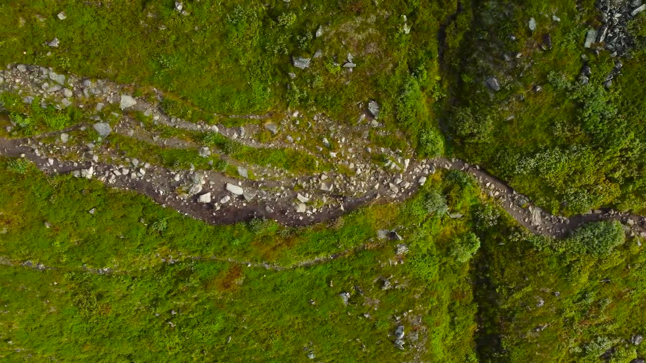 Top down aerial video of a person hiking and walking on green grassy and rocky Sweden terrain during a cloudy day. Filmed from high altitude and the footage follows the person walking down below.