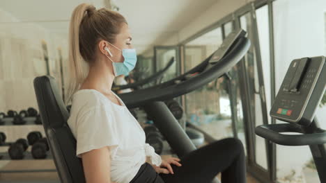 una joven atleta con mascarilla usa una máquina de ejercicios y bebe agua en el gimnasio