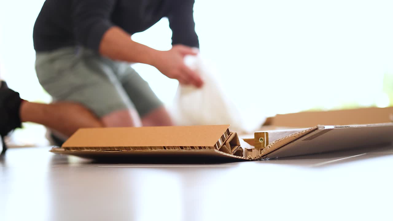 A person unpacks and assembles a wooden item from a box on a table in bright lighting