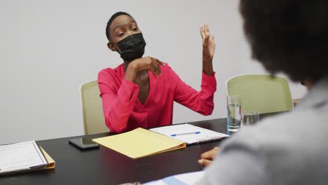 Mujer-De-Negocios-Afroamericana-Con-Mascarilla-Apuntando-A-La-Pantalla-Y-Hablando-Con-Colegas-En-La-Reunión