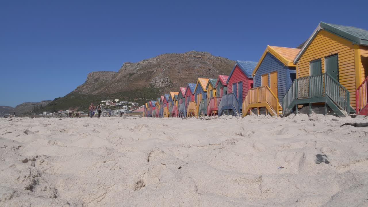 Dos mujeres caminan junto a las icónicas, vibrantes y multicolores casetas de playa en Muizenberg, Ciudad del Cabo, con una escarpada cordillera visible detrás de ellas bajo un cielo azul claro