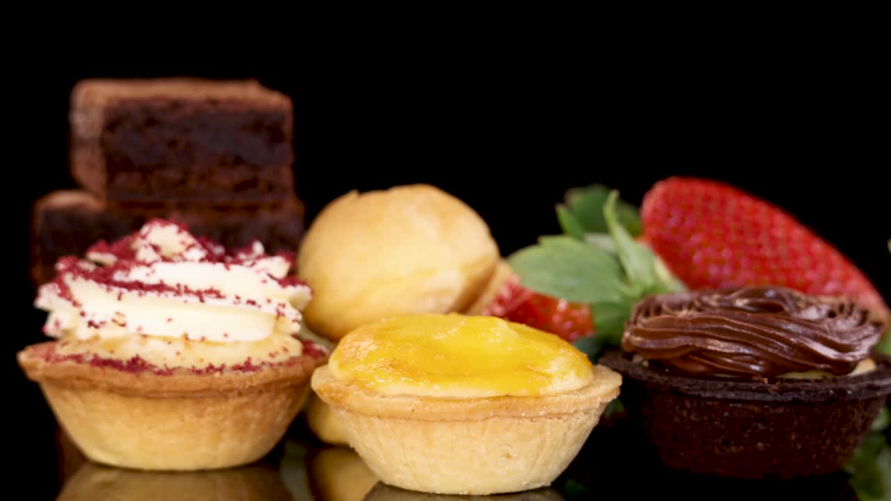 A hand reaches in to pick up a brownie from an elegant arrangement of assorted mini tarts, chocolate cream desserts, strawberries, and baked sweets on a glossy black surface with bright, even lighting