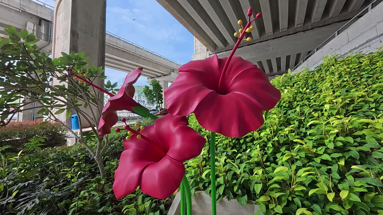 A large hibiscus sculpture with three blooming flowers stands prominently in a public park, adding a vibrant, artistic touch to the surroundings.