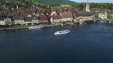 Aerial-orbit-of-touristic-boats-in-Rhine-river-next-to-Stein-am-Rhein-medieval-town-in-green-valley,-hills-in-background,-Switzerland