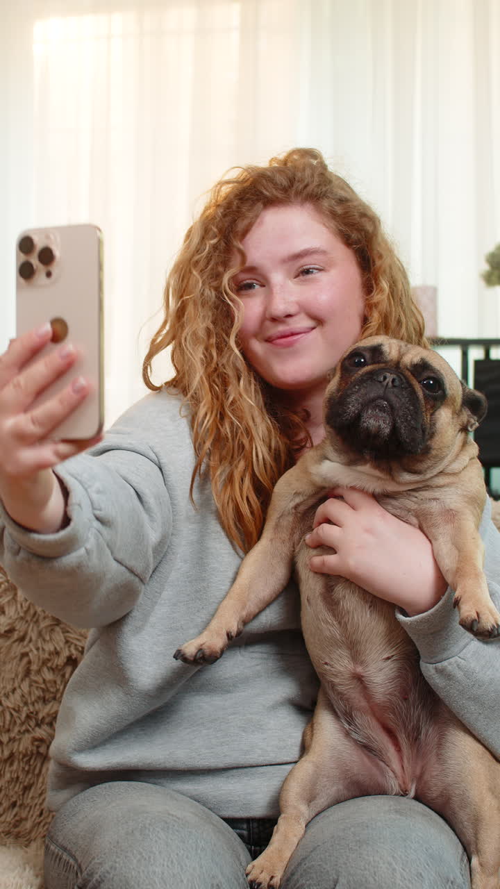 Young woman taking selfie with pug dog on home sofa smiling with joy while holding cute pet indoors